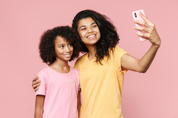 Smiling african american young woman and little kid girl sisters in casual t-shirts hugging doing selfie shot on mobile phone isolated on pastel pink background studio portrait. Family day concept.