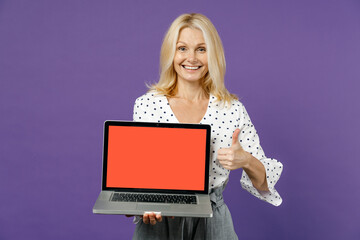 Cheerful elderly gray-haired blonde woman lady 40s 50s years old in white dotted blouse hold laptop pc computer with blank empty screen showing thumb up isolated on violet background studio portrait.