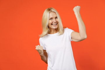 Cheerful happy elderly gray-haired blonde woman lady 40s 50s years old in white casual t-shirt standing doing winner gesture clenching fists isolated on bright orange color background studio portrait.
