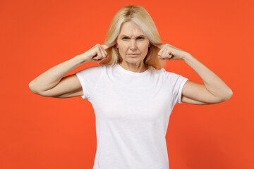 Dissatisfied elderly gray-haired blonde woman lady 40s 50s years old in white casual t-shirt stand covering ears with fingers looking camera isolated on bright orange color background studio portrait.