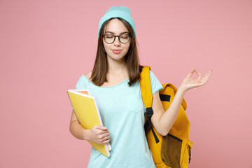 Young woman student in blue t-shirt hat glasses backpack hold notebooks hold hands in yoga gesture relaxing meditating isolated on pink background. Education in high school university college concept.