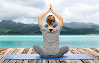 yoga, sport and healthy lifestyle concept - woman meditating in lotus pose on mat over ocean and mountains in french polynesia on background