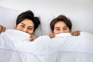 Happy Asian homosexual gay men male couple hiding under blanket in bed together. LGBT and sexual diversity concept. Looking at camera.