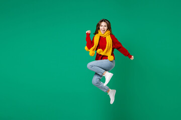 Full length side view happy joyful young brunette woman 20s in casual knitted red sweater yellow scarf jumping doing winner gesture looking camera isolated on green color background studio portrait.