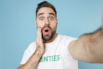 Close up of shocked young bearded man in volunteer t-shirt doing selfie shot on mobile phone put hand on cheek isolated on blue background. Voluntary free work assistance help charity grace concept.