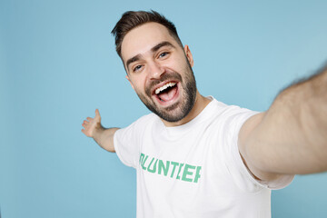 Naklejka premium Close up of excited young bearded man in volunteer t-shirt doing selfie shot on mobile phone pointing hand aside isolated on blue background. Voluntary free work assistance help charity grace concept.
