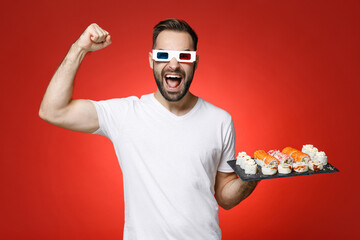 Joyful young man 20s in white t-shirt 3d glasses watching movie film showing biceps doing winner gesture hold makizushi sushi roll served on plate traditional japanese food isolated on red background.