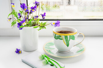 A cup of coffee, flowers of the field pansies, phone and headphones on the windowsill