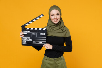 Smiling young arabian muslim woman in hijab black green clothes hold classic black film making clapperboard isolated on yellow color background studio portrait. People religious lifestyle concept.