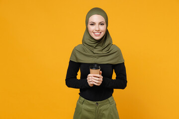 Smiling pretty young arabian muslim woman in hijab black green clothes hold paper cup of coffee or tea isolated on bright yellow color background, studio portrait. People religious lifestyle concept.