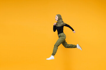 Fototapeta na wymiar Full length side view of cheerful young arabian muslim woman in hijab black green clothes jumping like running isolated on yellow color background studio portrait. People religious lifestyle concept.
