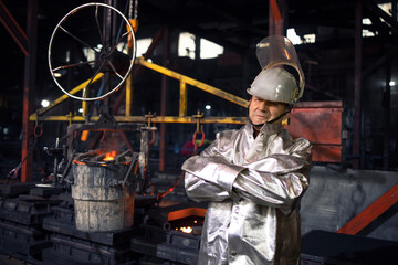 Fototapeta na wymiar Portrait of workman in aluminized high temperature protection suit with arms crossed standing in foundry steel production factory. In background bucket with liquid molten iron. 