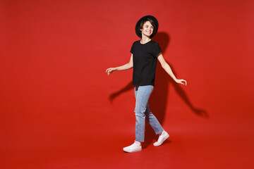 Fototapeta na wymiar Full length side view of smiling pretty cute young brunette woman 20s years old wearing casual basic black t-shirt hat standing looking aside isolated on bright red color background studio portrait.