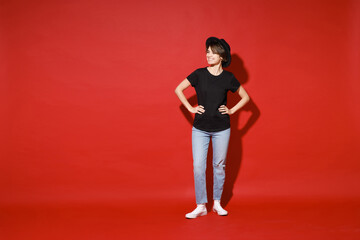 Full length of funny young brunette woman 20s years old wearing casual basic black t-shirt hat standing with arms akimbo on waist looking aside isolated on bright red color background studio portrait.