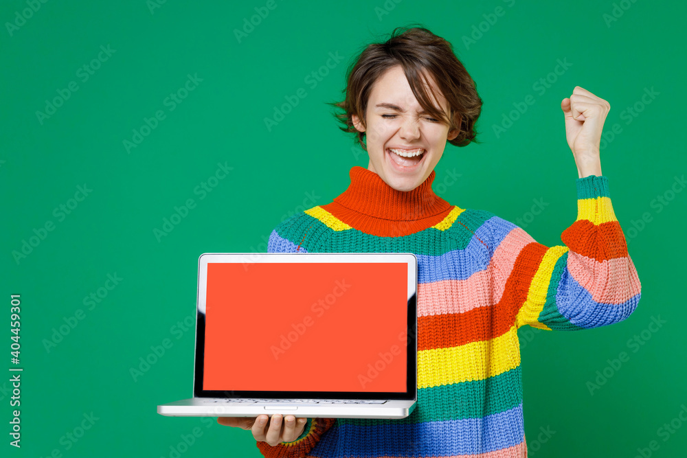 Canvas Prints Happy joyful young brunette woman 20s in basic casual colorful sweater hold laptop pc computer with blank empty screen doing winner gesture isolated on bright green color background studio portrait.