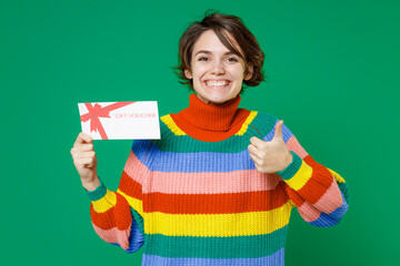 Smiling funny young brunette woman 20s years old in basic casual colorful sweater standing hold in hands gift certificate showing thumb up isolated on bright green color background studio portrait.