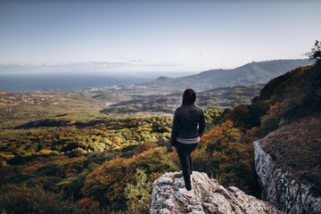 caucasian woman in dark jeans and a dark jacket stands on the edge of a cliff, and looks at the landscape of an autumn valley going to the sea against a blue sky.