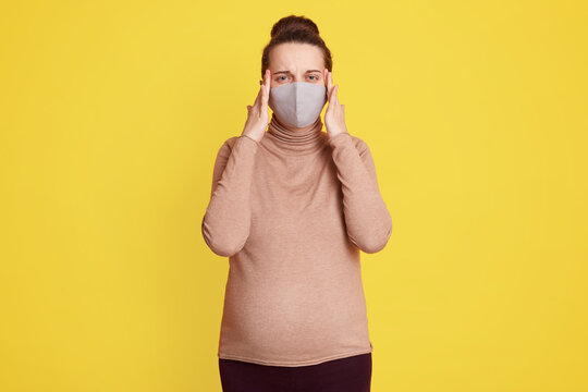 Beautiful pregnant woman in medical face mask keeping fingers on temples, suffering from headache, being scared to get infected of corona virus, wearing casually, posing against yellow wall.