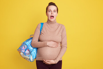 Shocked expectant mother posing isolated over yellow background with widely opened mouth, being very afraid to go to maternity home, keeping hands on her belly.