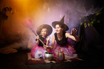 Beautiful brunette mother and cute little daughter looking as witches in special dresses and hats conjuring with a pot in room decorated for Halloween. Halloween style photo shoot.