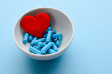 Blue medical capsules in white bowl and a red heart on a blue background. Concept of prevention and treatment of cardiovascular diseases, Valentine's day. Medical flat lay. World Pharmacist's Day