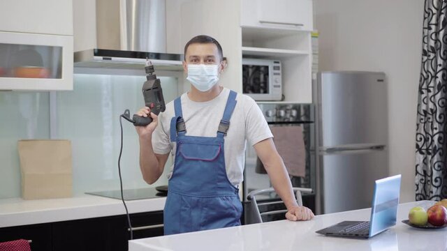 Positive Confident Repair Man In Covid-19 Face Mask Posing With Electric Tool In Apartment. Young Caucasian Worker Fixing Breakage On Coronavirus Pandemic. Professionalism Concept.