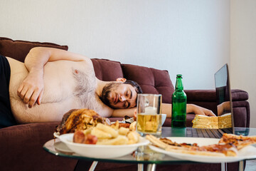 Drunk obese young man sleeping on sofa after party at home. Table with food and drinks alcohol is...