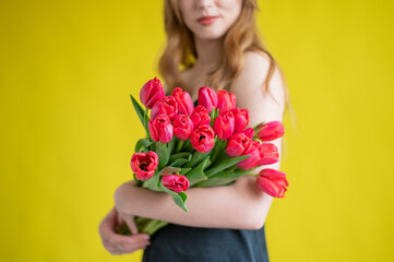 Unrecognizable woman with a bouquet of red tulips on a yellow background. A girl in a black dress holds an armful of flowers. A gift for International Women's Day. Spring holiday.