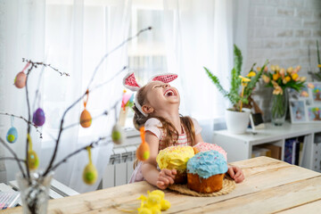 funny little girl with rabbit ears sitting at the table and holding Easter cakes