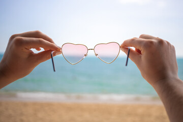 romantic holidays, vacation, love concept. hands holding pink heart-shaped sunglasses. in the background a sandy beach and blue ocean on a sunny day in blur
