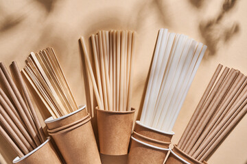 Renewable individual objects for home use, bamboo or paper straws, disposable cups and wooden coffee stirrers on beige with shade from the leaves on the beach. Zero waste. Top view.