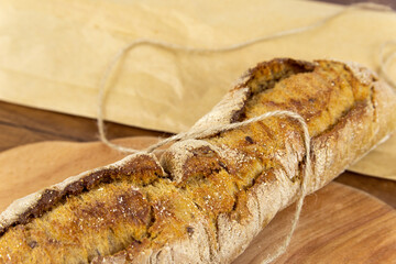 One baguette on a wooden cutting board, with a paper bag on the background, on a wooden background. A crispy white baguette lies on a dark wooden table, not all in focus. Close-up.