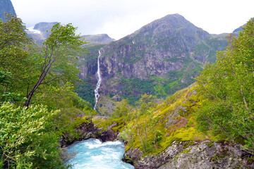 Pathway to Briksdal or Briksdalsbreen glacier in Olden. Landscape of Norway with huge mountains, waterfall and azure river. Windy and rainy weather.