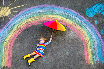 Happy little toddler girl in rubber boots with rainbow sun and clouds with rain painted with colorful chalks on ground or asphalt in summer. Cute child with umbrella having fun. creative leisure © Irina Schmidt