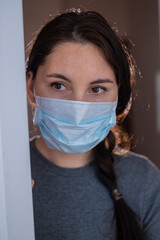 A beautiful young brunette woman in a medical mask looks out from behind the balcony door. Epidemic, quarantine, lockdown