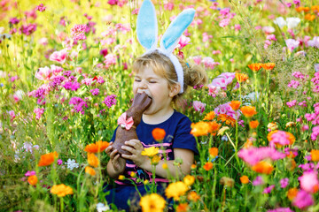 Portriat of adorable, charming toddler girl with Easter bunny ears eating chocolate bunny figure in flowers meadow. Smiling happy baby child on sunny day with colorful flowers, outdoors.