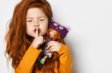 Cheerful small red haired girl in yellow comfortable longsleeve standing holding toy doll in hands...