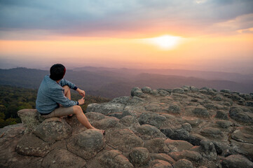 Achievement concept. Asian man raising hands sitting on Nodule rock field called Lan Hin Pum enjoy looking at sunset over scenic landscape of Phu Hin Rong Kla National Park in Phitsanulok, Thailand.