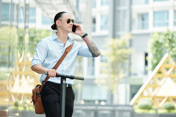 Positive stylish young man standing on electric scooter and talking on phone with colleague