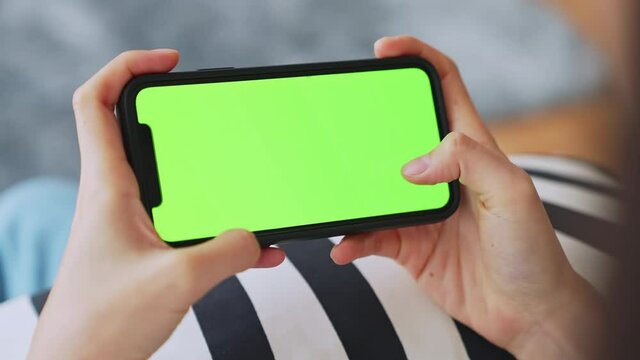 Young Asian woman holding in hands a mock up smartphone with green screen for playing games, chromakey screen for advertising. the living room at home.