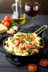 Small pan with fettuccine and sea food decorated with greens and cherry tomatoes, black napkin, a glass of wine, bottle with olive oil around on the black wooden table. Close up