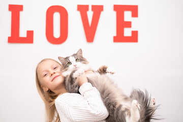 A girl in a white sweater plays with a gray cat against the background of the red inscription LOVE. Greeting card with a child for Valentine's Day, Mother's Day, Birthday