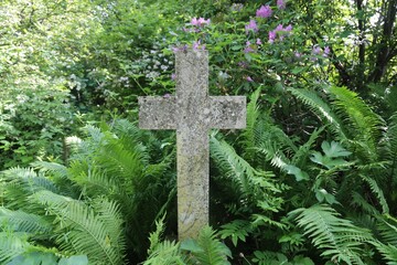 A lonely, isolated, plain stone cross situated in an overgrown garden of ferns, shrubs and trees.