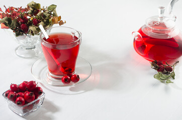 Autumn red herbal tea in transparent teapot, cup with dry leaves, rose hip, hawthorn berries in sunlight on white wood board, copy space.