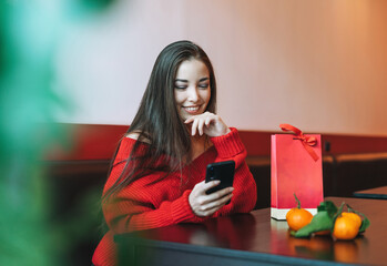 Beautiful smiling young asian woman in red clothes using mobile with gift bag with mandarins in the cafe, celebrating Chinese New Year