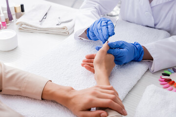 partial view of manicurist in latex gloves using cuticle pusher while making manicure to client