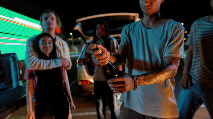 A group of well-dressed friends waiting for a young tattooed guy to open a bottle of champagne during a party with a car and a big led screen on a background