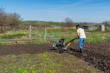 Man in wellingtons with cultivator ploughs ground in sunny day. Land cultivation, soil tillage. Spring work in garden. Gardening concept