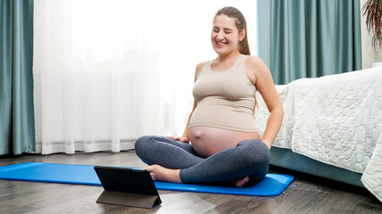 Beautiful pregnant woman talking to her yoga teacher via tablet computer. Concept of healthy lifestyle, healthcare and sports during pregnancy