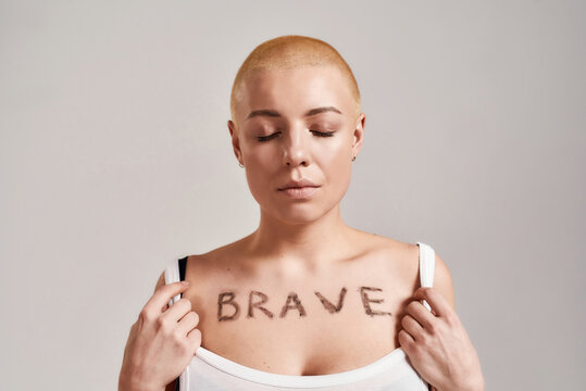 Portrait of a beautiful young caucasian woman showing her chest with the word Brave painted on it while posing with eyes closed isolated over grey background
